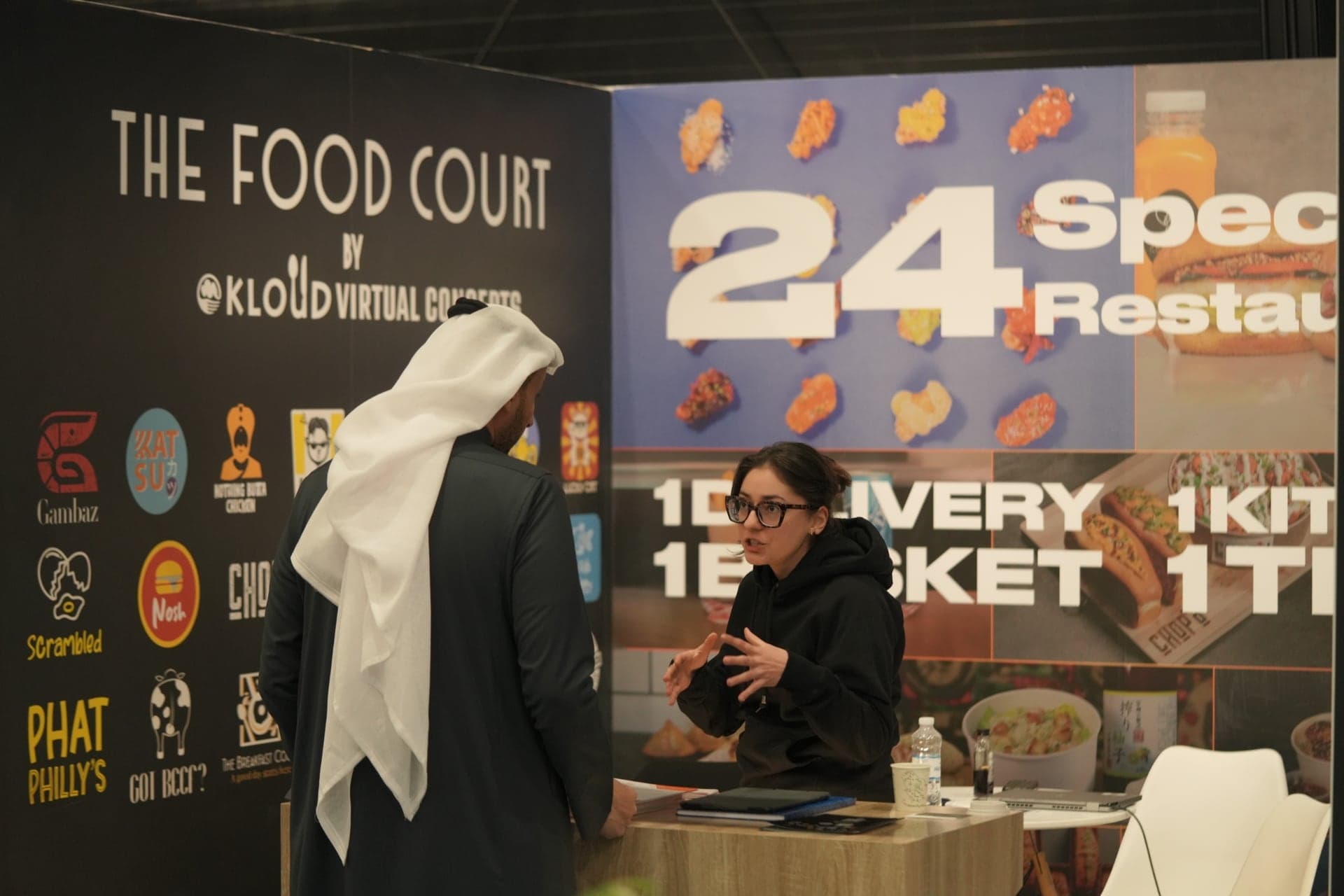 An exhibitor explaining a multi-brand food court concept to a visitor at her stand
